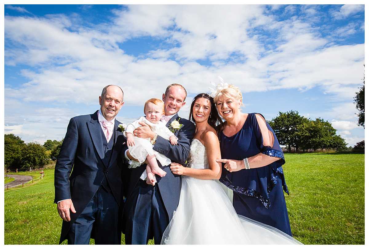Family photos - bride and groom, holding their baby girl with brides mother and step day. Smiling, all nice and close with their heads with a beautiful blue sky