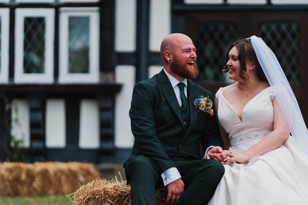 Bride and Groom giggling outside Wistanstow Village Hall sat on a hay bale giggling between themselves / Shropshire Wedding Photographer