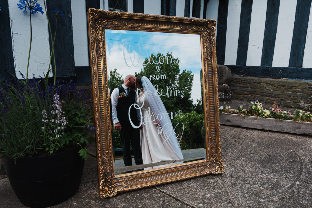 Bride and Groom kissing in the mirror with welcome not outside Wistanstow Village Hall, Craven Arms
