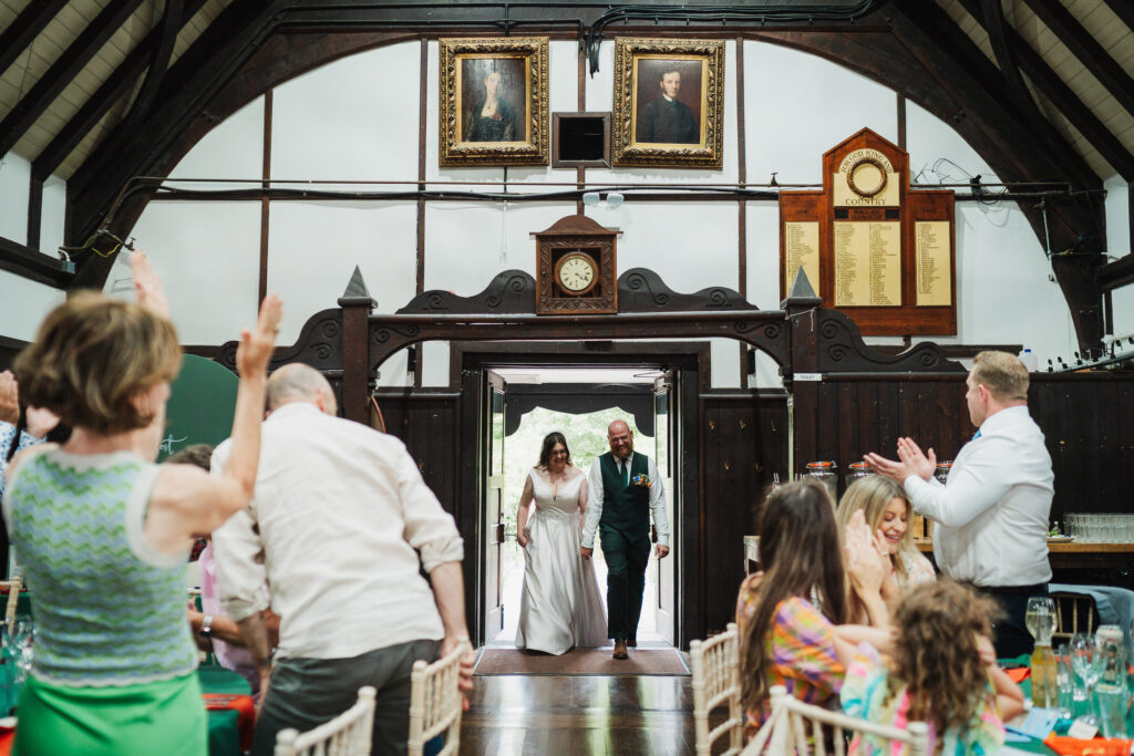 Guests applauding bride and groom as they enter their wedding reception at Wistanstow Village Hall / Charlotte Giddings Photography