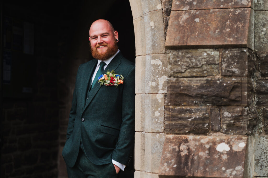 Groom dressed in green three piece suit outside Wistanstow Village Hall and floral decoration by Shropshire Flower Company
