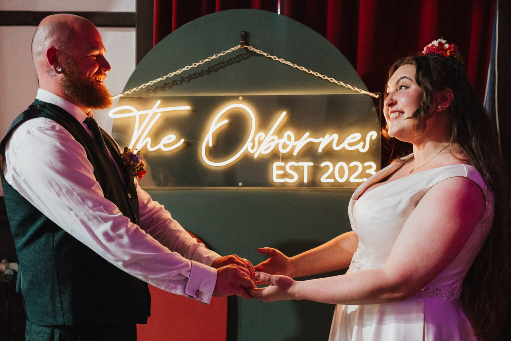 Bride and Groom holding hands in front of the neon sign with their names at Wistanstow Village Hall, Craven Arms
