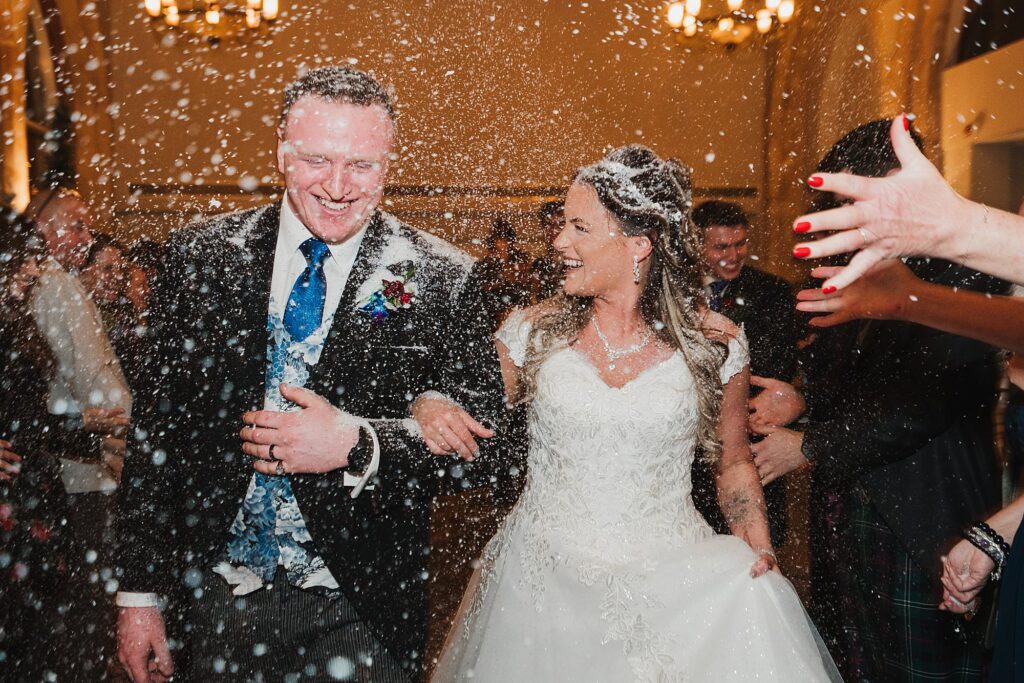 Bride and Groom covered in fake snow as they walk through the confetti tunnel at the Bolton Wedding