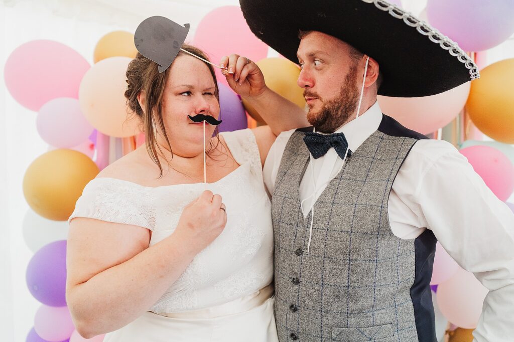 Bride and Groom partaking in the photo booth fun at a DIY wedding and pulling funny expressions with props // Shropshire Wedding Photographer