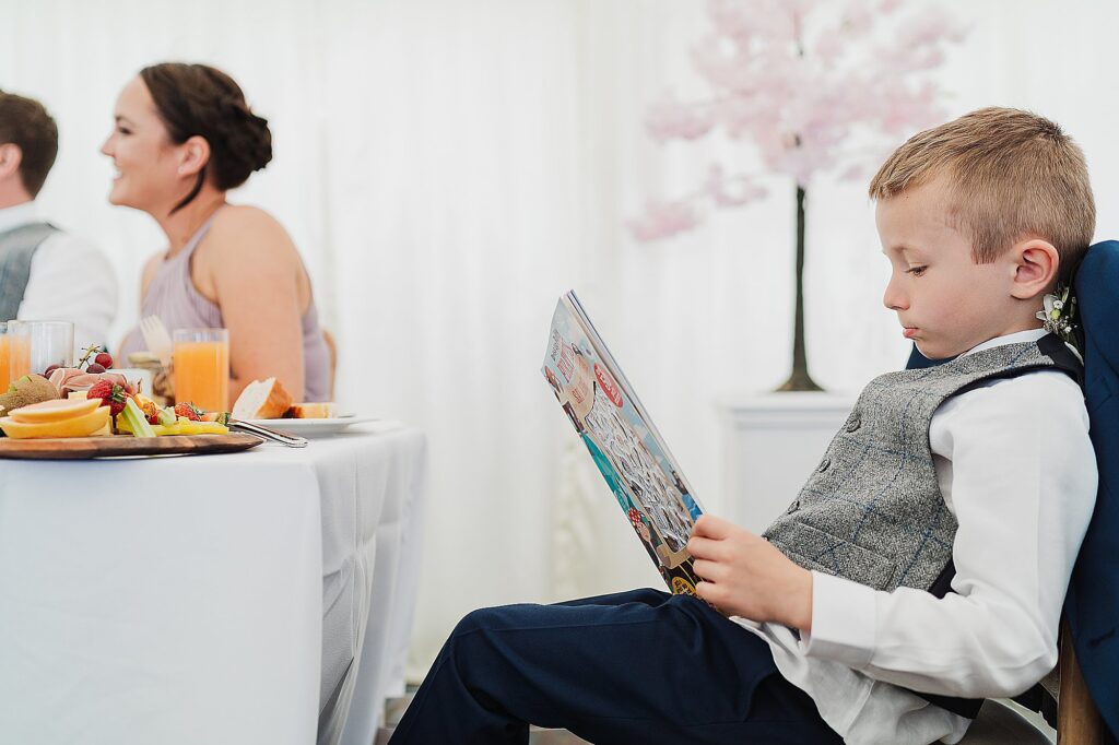 Young Boy sitting amongst the guests readying a comic and enjoying some escape at a wedding // Charlotte Giddings Photography