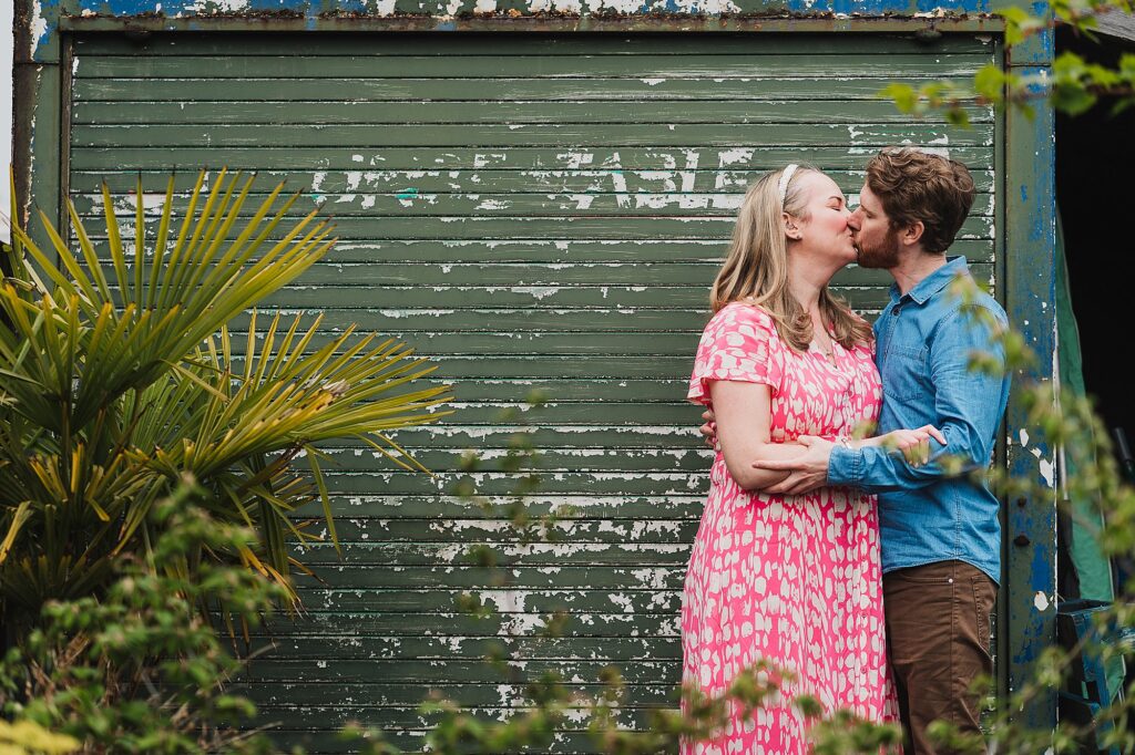 Bride and Groom to be kissing against a green metal shutter door amongst lots of green foliage // Pre Wedding Shoot Shropshire