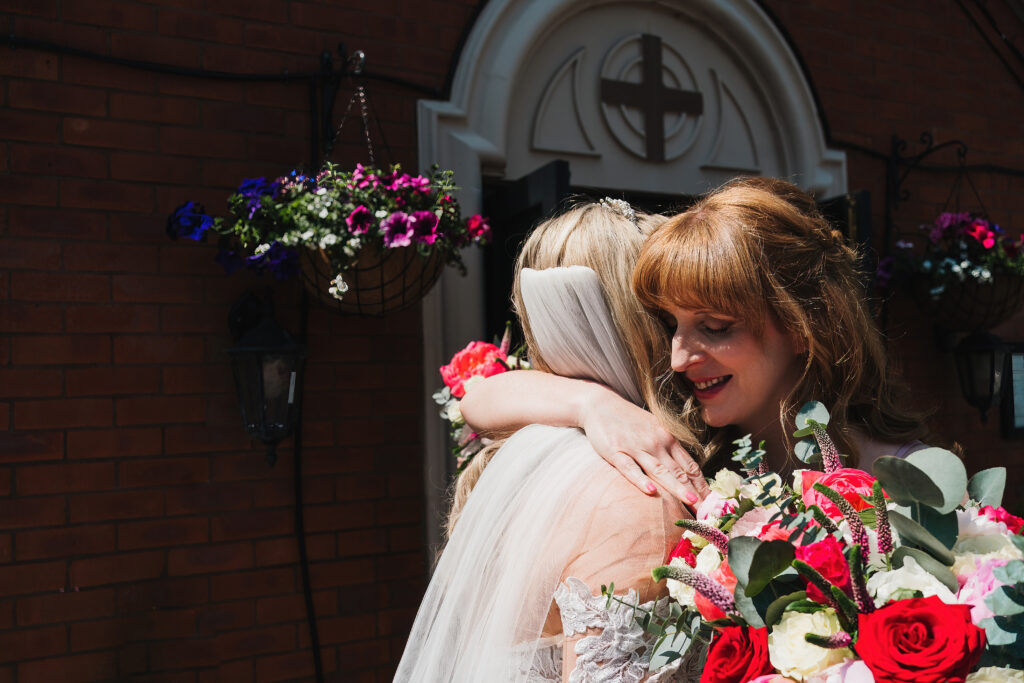 Bridesmaid embracing the bride outside a Cathelic Church, Nantwich in beautiful sunshine // Shropshire Wedding Photographer