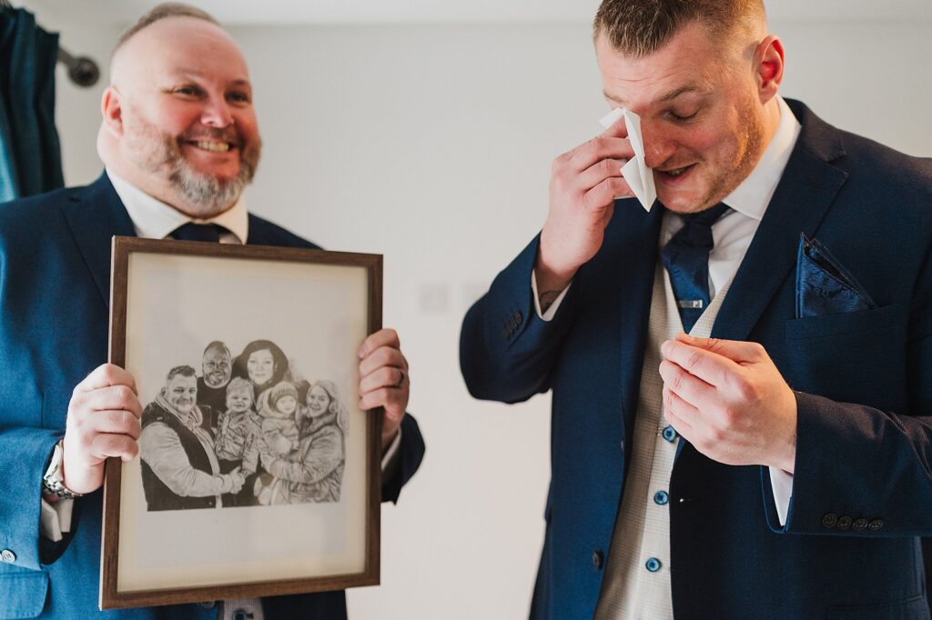 Groom wiping away tears after receiving a drawing of his mum who is no longer with us with all the family // Old Palace Wedding Photographer