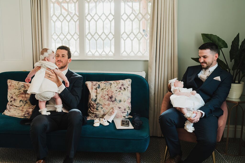 Two men in similar suits, holding their baby daughters in similar outfits having some time out in a quiet area at a Combermere Abbey Wedding // Shropshire Wedding Venue
