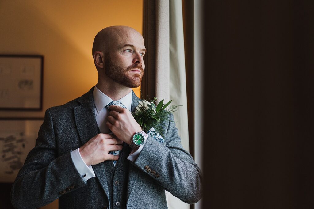Groom adjusting his tie and looking out the window at Hill Valley Hotel Whitchurch, Shropshire // Charlotte Giddings Photography