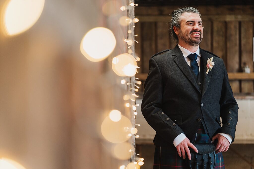 Groom looking at his guests as they arrive at his Grange Barn Wedding Cheshire with twinkly lights in the foreground // Charlotte Giddings Photography