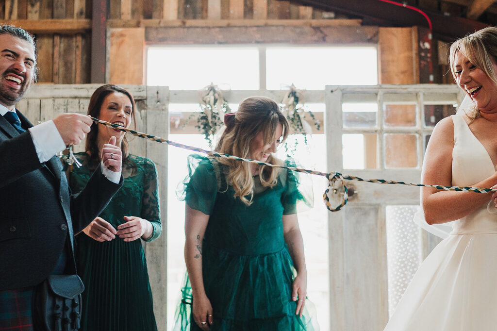 Bride and Groom literally tying the knot during the hand fasting ceremony with both sisters assisting at Grange Barn wedding venue Cheshire