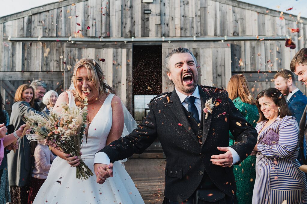 Bride and groom crying with laughter as wedding guests throw confetti on them with Grange Barn in the Background // Charlotte Giddings Photography