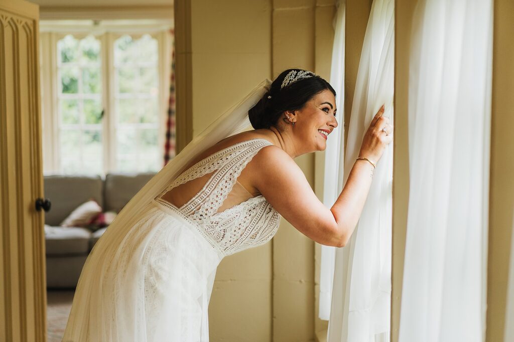 Bride, with a big bursting smile peeking through the curtains in the Stone Lodge at Combermere Abbey