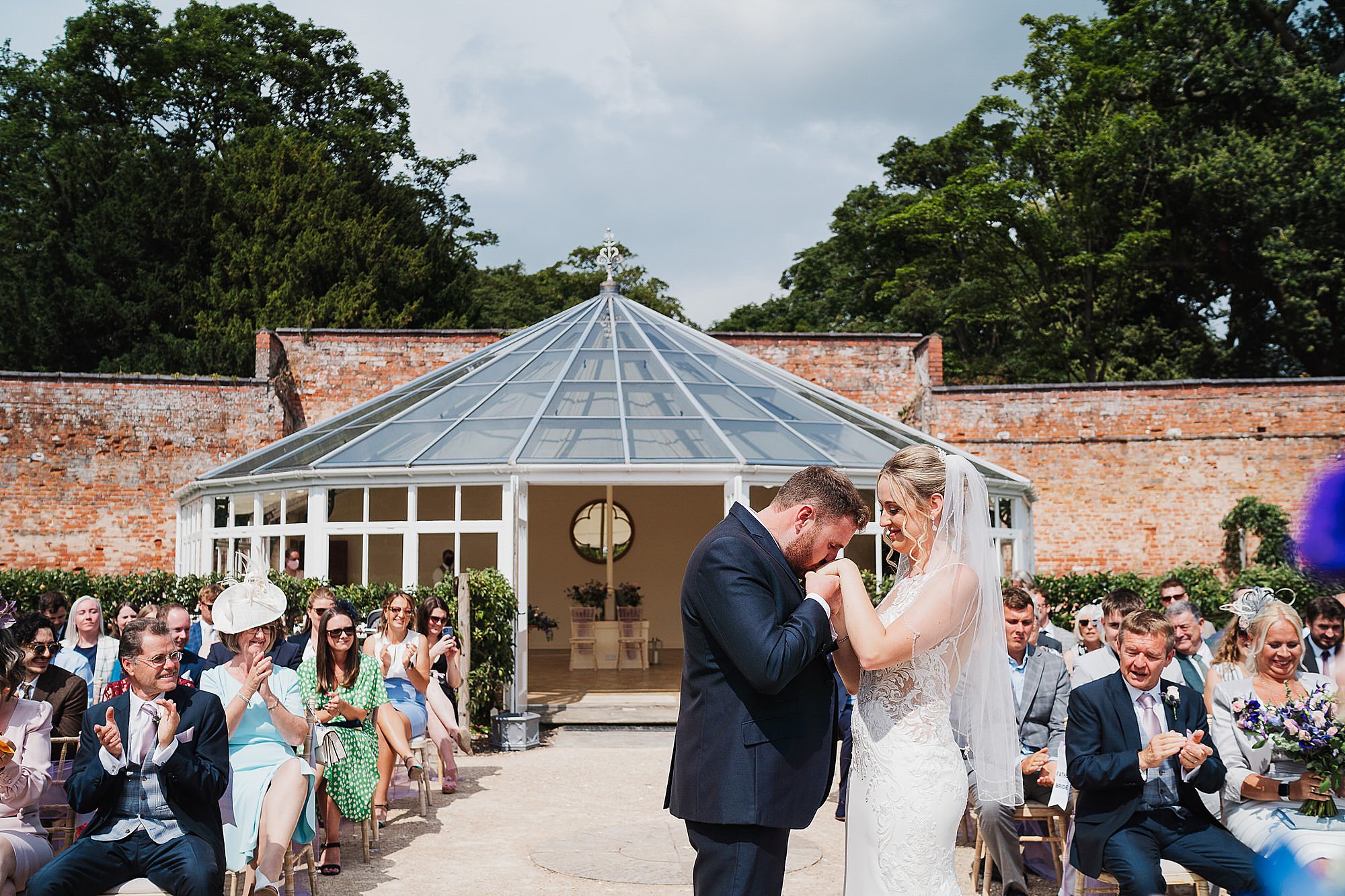 Groom kissing brides hand at end of ceremony outside Glasshouse, Combermere Abbey Wedding Venue